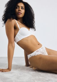 Soutien-gorge blanc rembourré avec des bretelles fines et un ensemble de culotte en dentelle assorti avec des motifs floraux et des bords festonnés, sur un fond clair.