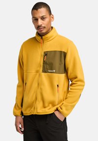Gelbe Fleece-Jacke mit grüner Brusttasche. Verfügt über einen hohen Kragen, zwei seitliche Reißverschlusstaschen und Reißverschlussdetails in Orange.
