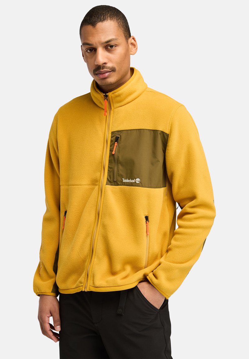 Gelbe Fleece-Jacke mit grüner Brusttasche. Verfügt über einen hohen Kragen, zwei seitliche Reißverschlusstaschen und Reißverschlussdetails in Orange.
