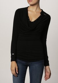 Vrouw in een zwarte top met lange mouwen en gedrapeerde halslijn met metalen oogjes, gecombineerd met donkerblauwe jeans, staande tegen een effen achtergrond.