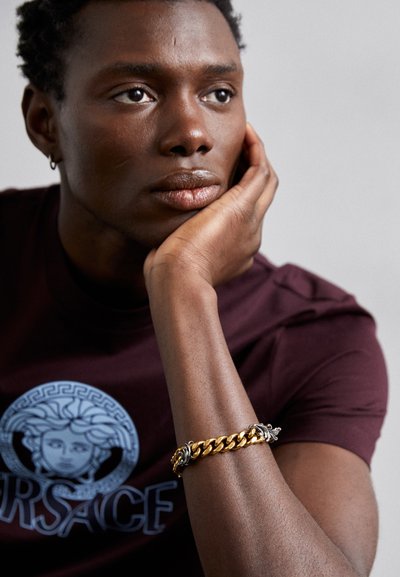 Burgunderfarbenes T-Shirt mit einem grauen Grafiken eines Gesichts, kombiniert mit einem massiven Gold- und Silberschmuckarmband am Handgelenk eines Models.