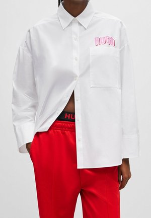 Personne portant une chemise blanche à boutons avec le texte rose "HUGO" sur la poche, un pantalon rouge et une ceinture noire avec le logo rouge "HUGO" visible.