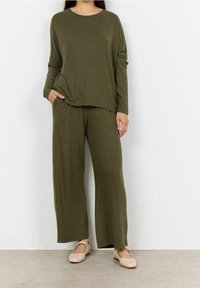 Frau in olivgrünem Langarmoberteil und weiten Hosen mit beigen Flats, stehend vor einer weißen Wand.