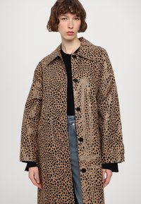 Brown, oversized coat with black and silver leopard print, classic collar, button closure, and long sleeves. Worn over a black top.