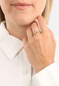 Anello in tono oro con design a torsione, caratterizzato da un piccolo diamante decorativo. Indossato su una mano contro una camicia chiara con colletto.