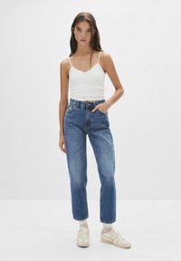 Lichte witte tanktop met dunne schouderbandjes, gecombineerd met hooggetsneden blauwe denim jeans. Model draagt crèmewitte sneakers en staat tegen een lichte achtergrond.