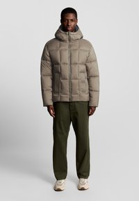 Pufferjack in lichtbeige met een capuchon, gewatteerd ontwerp en een ritssluiting aan de voorkant. Draag het met olijfgroene broeken en beige sneakers.