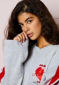 Siva majica z dolgimi rokavi, z rdečimi poudarki in grafiko srca. Tkanina se zdi mehka s vidnimi šivi.