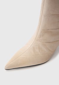 Stivale beige a punta realizzato in pelle testurizzata con motivo coccodrillo, caratterizzato da una silhouette elegante e una suola nera.