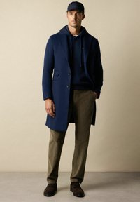 Granatowy wełniany płaszcz na granatowej bluzie z kapturem, zestawiony z oliwkowymi sztruksowymi spodniami i brązowymi zamszowymi butami. Detale obejmują kieszenie i zapięcie na guziki z przodu.