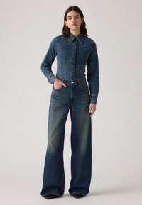 Chemise en denim avec un col et des manches longues, associée à un jean en denim à jambes larges. Les deux sont d'un bleu médium clair avec une finition légèrement délavée.