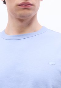 Νεαρό άτομο που φοράει γαλάζιο μπλουζάκι με λαιμόκοψη, με μικρό, αντίστοιχο μπλε λογότυπο στο αριστερό στήθος, σε απλό φόντο.