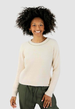 Donna con capelli ricci che sorride, indossa un maglione morbido color crema e pantaloni verde oliva con coulisse, in piedi davanti a uno sfondo chiaro e uniforme.