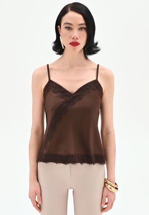 Femme aux cheveux courts noirs portant un caraco marron bordé de dentelle et un pantalon beige, accessoirisée de grandes boucles d'oreilles dorées et de bracelets.