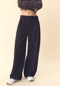 Czarne spodnie o szerokich nogawkach z gładką teksturą, zapinane z przodu na guziki i z zakładkami, zestawione z białymi butami sportowymi.