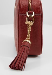 Sac à main en cuir rouge avec une finition texturée, quincaillerie dorée, accent en chaîne et un détail de pompon attaché à la tirette de la fermeture éclair.