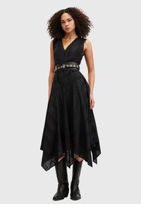 Mujer con pelo rizado que lleva un vestido negro sin mangas con dobladillo en pico, un cinturón negro ancho con ojetes dorados y botas negras hasta la rodilla.