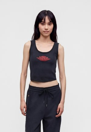Junge Frau mit langen dunklen Haaren, trägt ein schwarzes, bauchfreies Tanktop mit rotem Air Jordan-Logo und schwarze Hose mit Kordelzug, steht vor einem einfarbigen Hintergrund.