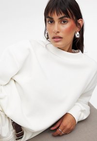 Biały, oversize'owy sweter z wysokim kołnierzem, ściągaczami oraz miękką teksturą. Modelka nosi srebrne kolczyki w kształcie kół, prezentując swobodny krój.