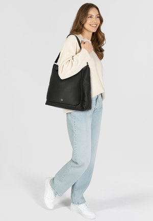 Borsa a spalla in pelle nera con una parte superiore curvata, cerniera argentata e logo. Il modello la tiene vicino a un maglione chiaro e jeans chiari.