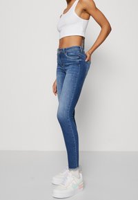Mujer que lleva jeans ajustados de tiro medio en azul, una camiseta corta blanca y zapatillas blancas con acentos en tonos pastel, de pie frente a un fondo liso.