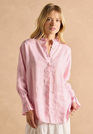 Young woman wearing loose pink and white vertical striped button-up shirt with ruffled collar and white skirt against beige background.