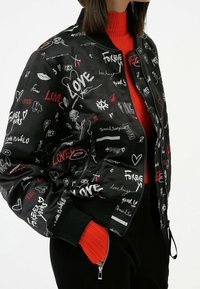 Giacca bomber nera con vari disegni e motivi bianchi e rossi scritti a mano, polsini a coste e chiusura con zip. Indossata sopra una maglietta rossa.