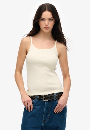 Prążkowany kremowy top na ramiączkach z cienkimi paskami i okrągłym dekoltem, noszony z niebieską spódnicą dżinsową i ozdobnym czarnym paskiem.