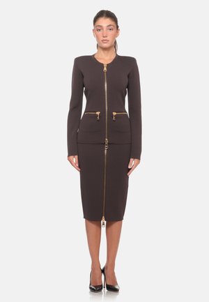 Donna in piedi che indossa un tailleur gonna aderente marrone scuro con cerniere dorate davanti e scarpe nere con tacco alto, su sfondo bianco semplice.