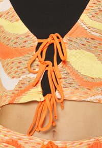 Top cropped tricotado em laranja, amarelo e branco com uma textura padronizada, apresentando laços ajustáveis na parte da frente e uma camada inferior preta.