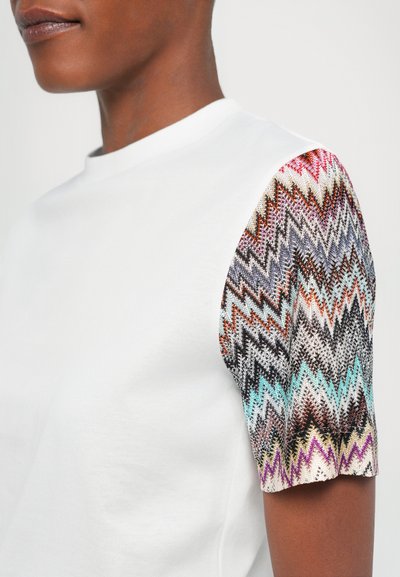 Camiseta blanca de algodón con un patrón de zigzag tejido en tonos multicolores en las mangas cortas. Cuello redondo clásico.