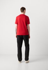 Rød kortærmet t-shirt lavet af bomuld, med rund hals, parret med sort denim jeans og hvide sko. Synlig tatovering på armen.