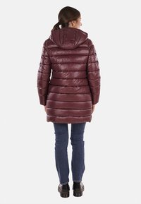 Giacca imbottita bordeaux con cappuccio, cuciture orizzontali e una texture leggermente lucida, abbinata a jeans scuri e calzature nere.