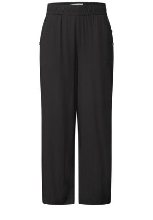 Femme debout portant un haut blanc à manches courtes, un pantalon noir ample, des chaussures beiges sans lacets et tenant un sac épaule beige.