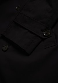 Detail černého rukávu kabátu s páskem a tmavými knoflíky, vystavující šití a texturu látky.