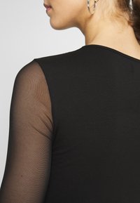 Svart topp med en solid tygdel på kroppen och genomskinliga nätärmar. Urringningen är rund, och det finns subtila texturdetaile på axlarna.