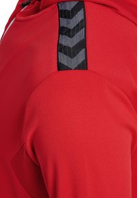 Rotes Sportshirt aus strukturiertem Stoff mit gebogenem Design und schwarzen Chevron-Musterakzenten an der Schulter.