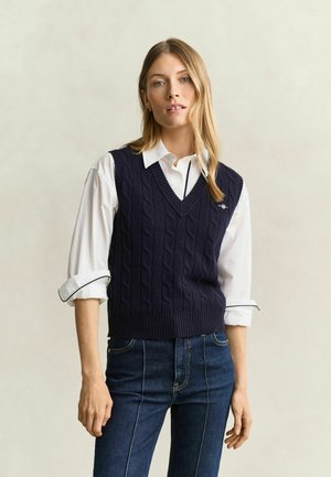 Femme portant un gilet en maille torsadée bleu marine par-dessus une chemise blanche à col avec manches retroussées et un jean bleu foncé, debout devant un fond uni.