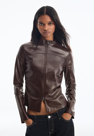 Femme portant une veste en cuir marron cintrée, les mains dans les poches, avec de longs cheveux foncés sur fond blanc.
