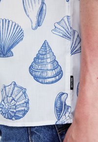Košile s krátkým rukávem s bílým pozadím a modrými vzory mořských mušlí, včetně hřebenatek a mušli, a se štítkem na boku.