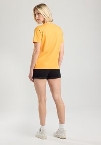 T-shirt en coton orange à manches courtes, vue de dos. Associé à un short en denim noir et des baskets blanches, avec des poignets retroussés et des chaussettes blanches.