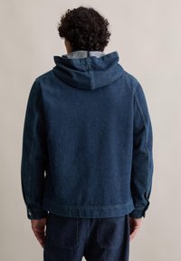 Blaue Denimjacke mit Kapuze, geradem Schnitt, langen Ärmeln und Knopfverschlussdetails. Der Stoff hat eine robuste und glatte Textur.