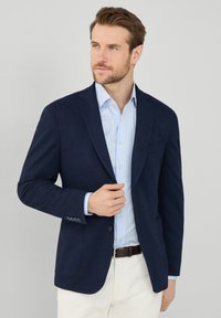 Blazer navy con rever a punta, chiusura a un bottone e due tasche frontali, indossato sopra una camicia a righe blu chiaro. Tessuto dalla consistenza liscia.