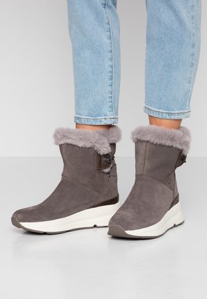 Stivali in suede grigi per l'inverno con bordo in pelliccia e dettaglio con bottone, indossati con jeans azzurri arrotolati, in piedi su un pavimento chiaro.