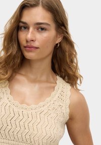 Top de tirantes de ganchillo beige con escote en puntilla, patrón en zigzag y un acabado texturizado. La modelo tiene el pelo suelto y ondulado y lleva pendientes pequeños de aro.