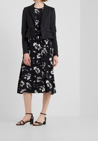 Žena v černém zkráceném sakou přes černé midi šaty s bílým květinovým vzorem, doplněné černými sandály na blokovém podpatku s řemínkem kolem kotníku.