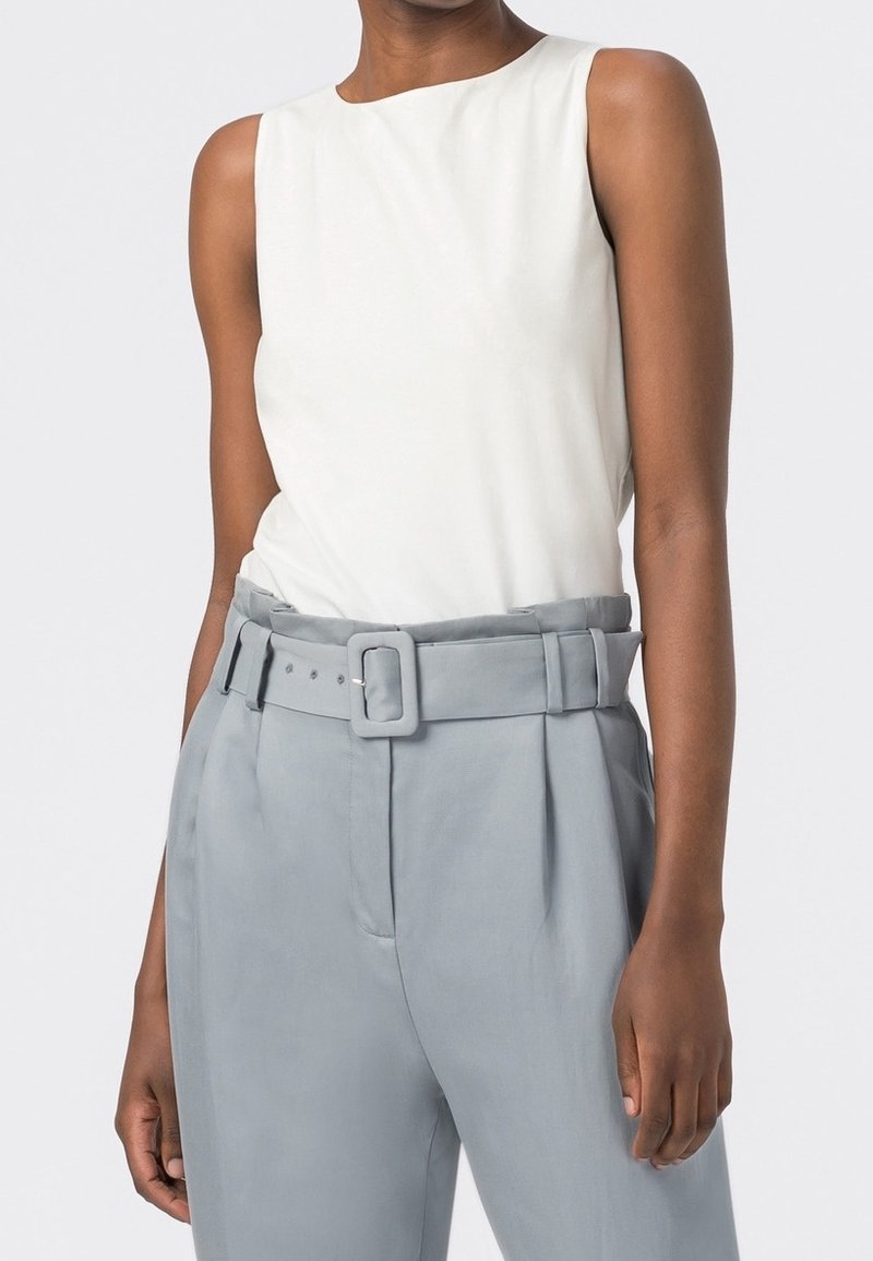 Vit ärmlös topp med rund halsringning, tillsammans med ljusgrå högmidjade byxor med bälte och sidofållar.