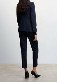 Costume bleu marine sur mesure avec blazer à un bouton et pantalon ajusté, associé à des escarpins noirs à talons aiguilles. Texture lisse et coupe élégante.