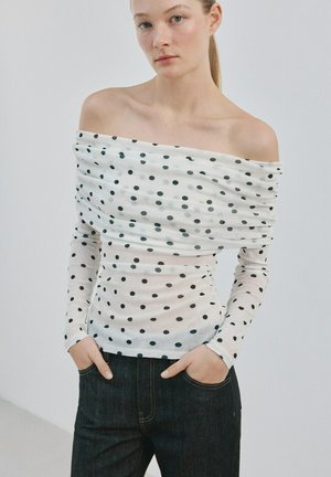 Tânără purtând un top alb cu buline negre, cu umerii goi, și blugi închiși la culoare, cu mâinile în buzunare, stând în picioare pe un fundal simplu.