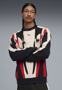 Uomo con capelli intrecciati e occhiali indossa una felpa Puma rossa, nera e bianca con le braccia incrociate davanti a uno sfondo grigio.